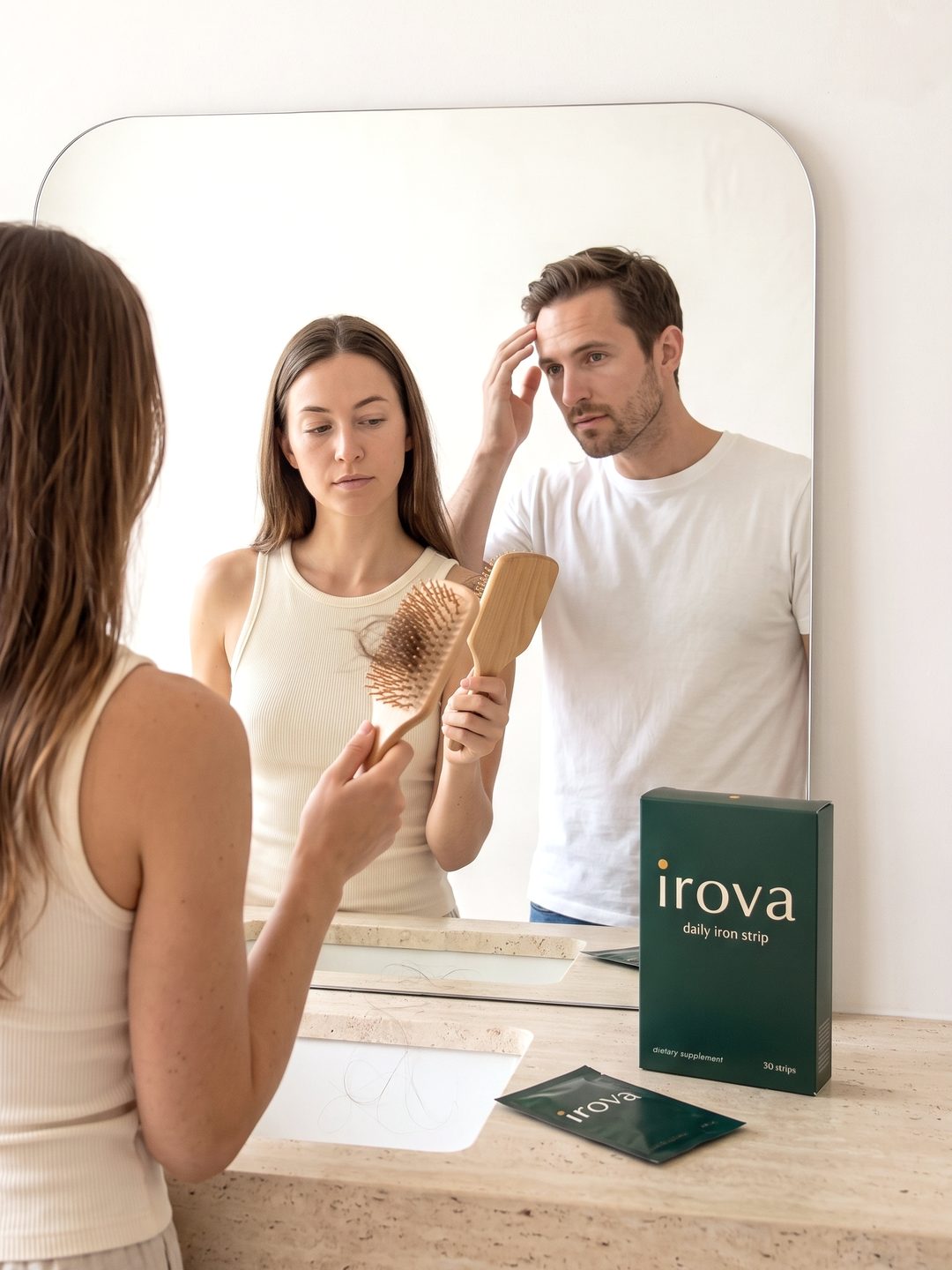 A woman and a man at a bathroom mirror, she holds a hairbrush with shed hair while he checks his hairline, with an Irova box on the vanity